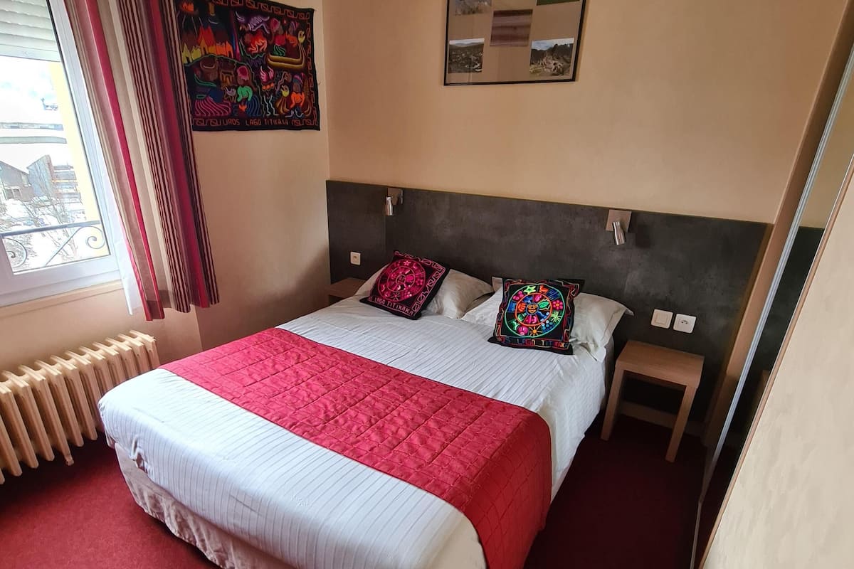 A hotel room with a large bed, red carpet, a radiator, a small table, and a wall with a framed picture.