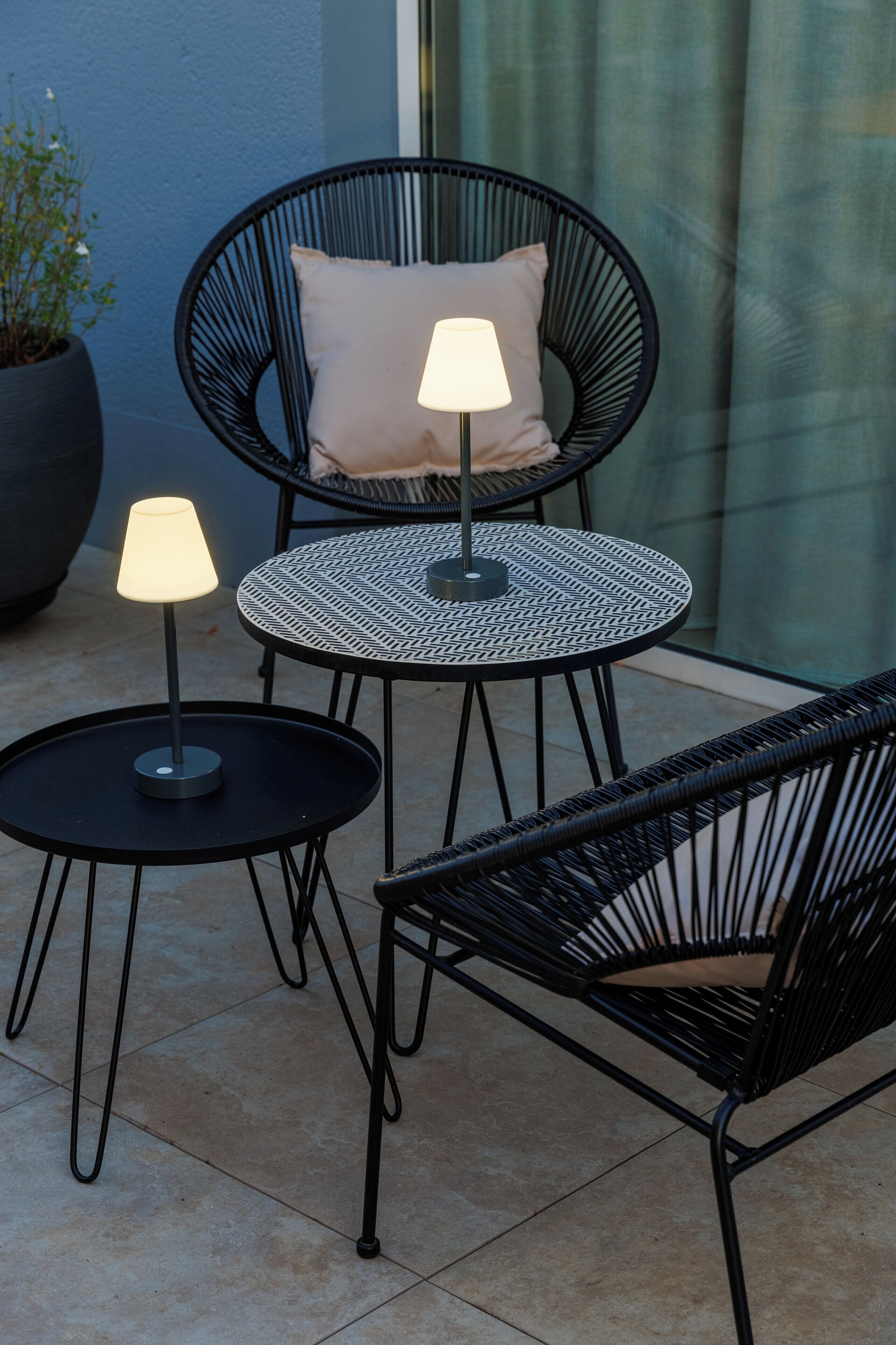 A patio area with two round tables, each with a black metal frame and a single table lamp. There is a black wicker chair with a cushion and a potted plant to the left.