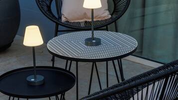 A patio area with two round tables, each with a black metal frame and a single table lamp. There is a black wicker chair with a cushion and a potted plant to the left.