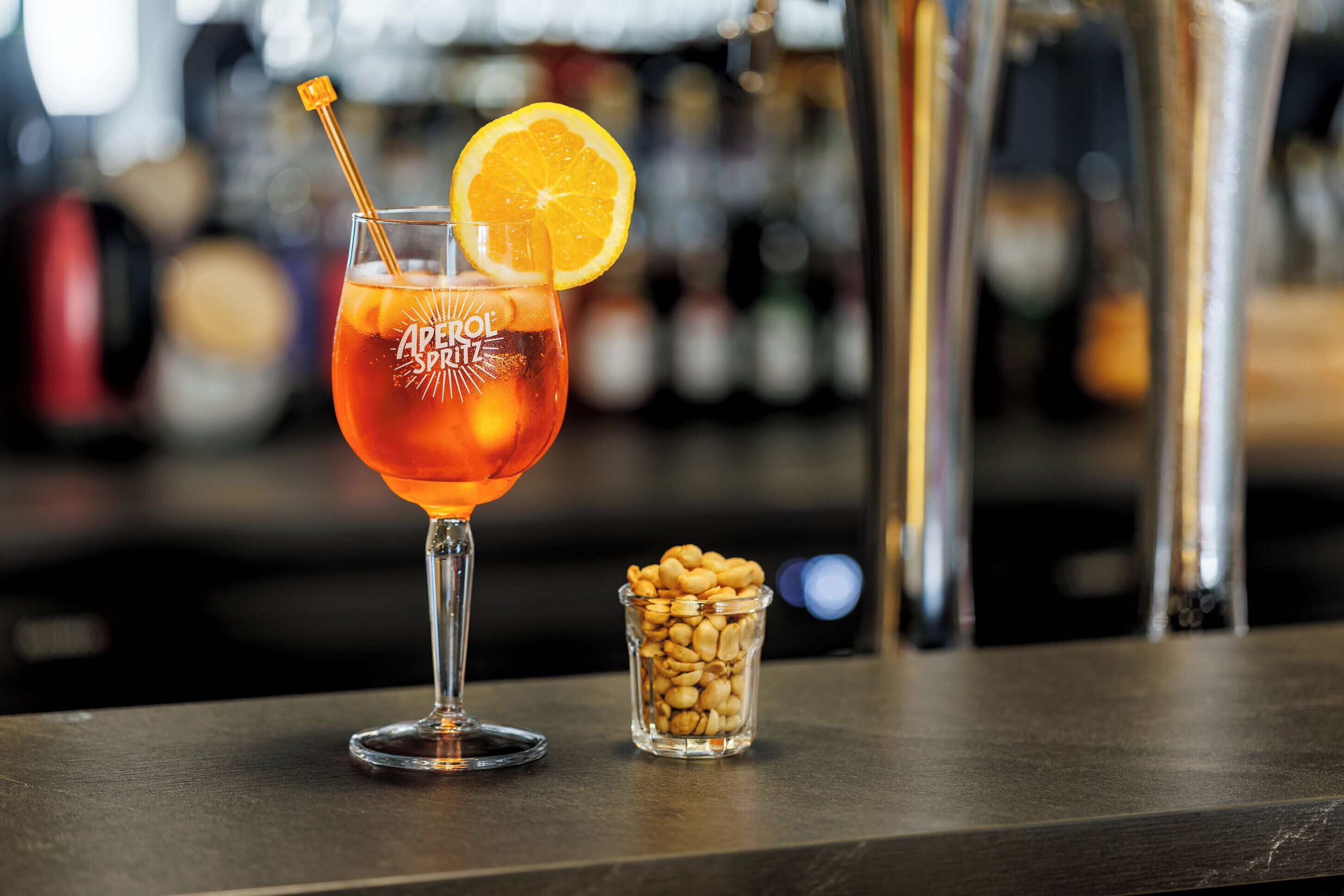 Una copa de cóctel con una rodaja de limón y una pajita, etiquetada como "Aperol Spritz", junto a un vaso pequeño de cacahuates sobre una barra.