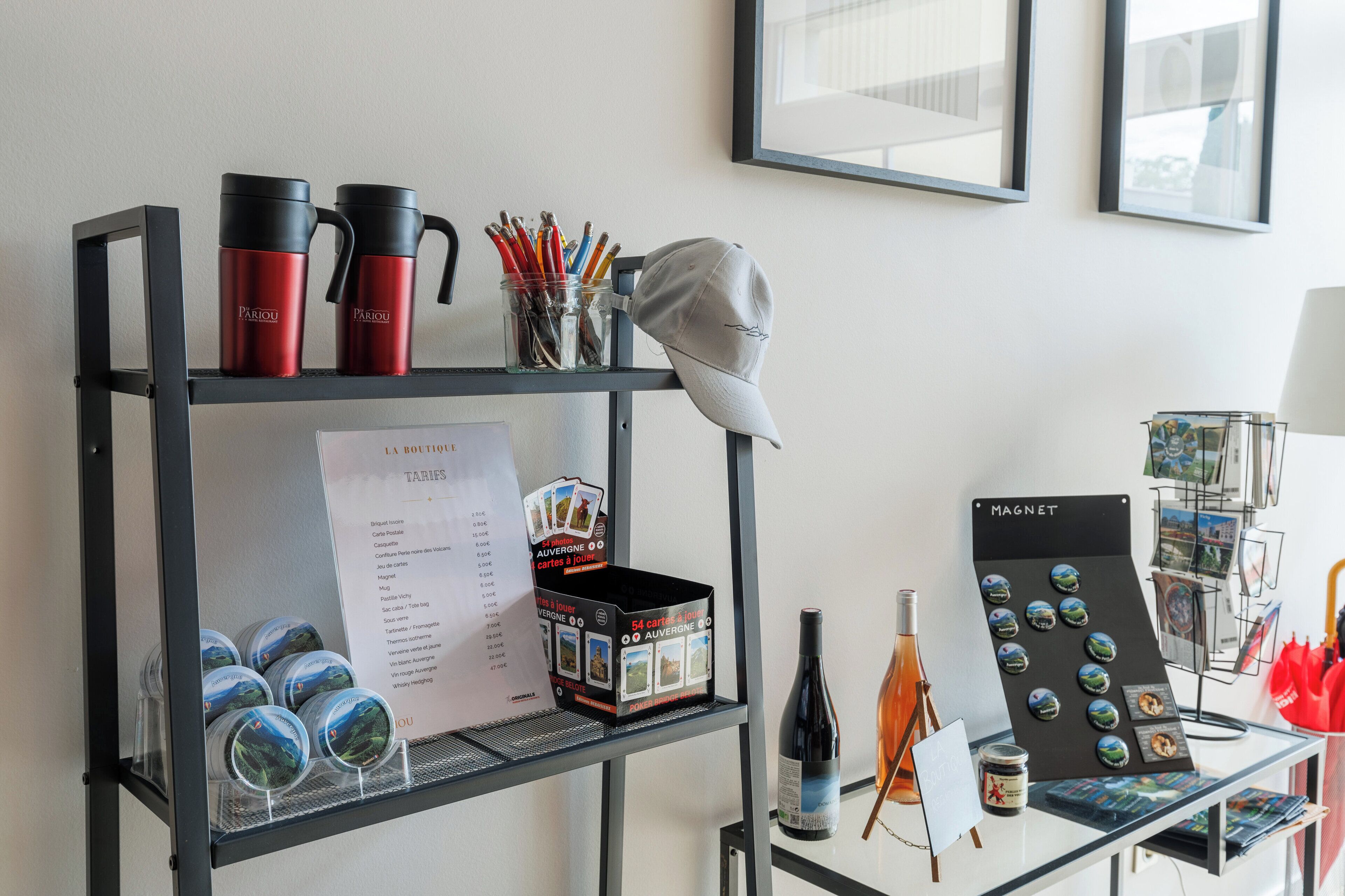 A metal shelf with red and black thermos flasks, a cap, and a jar of pencils. Below, there are tins of food and a wine bottle. A sign lists room rates, and a display of magnets is on the right.