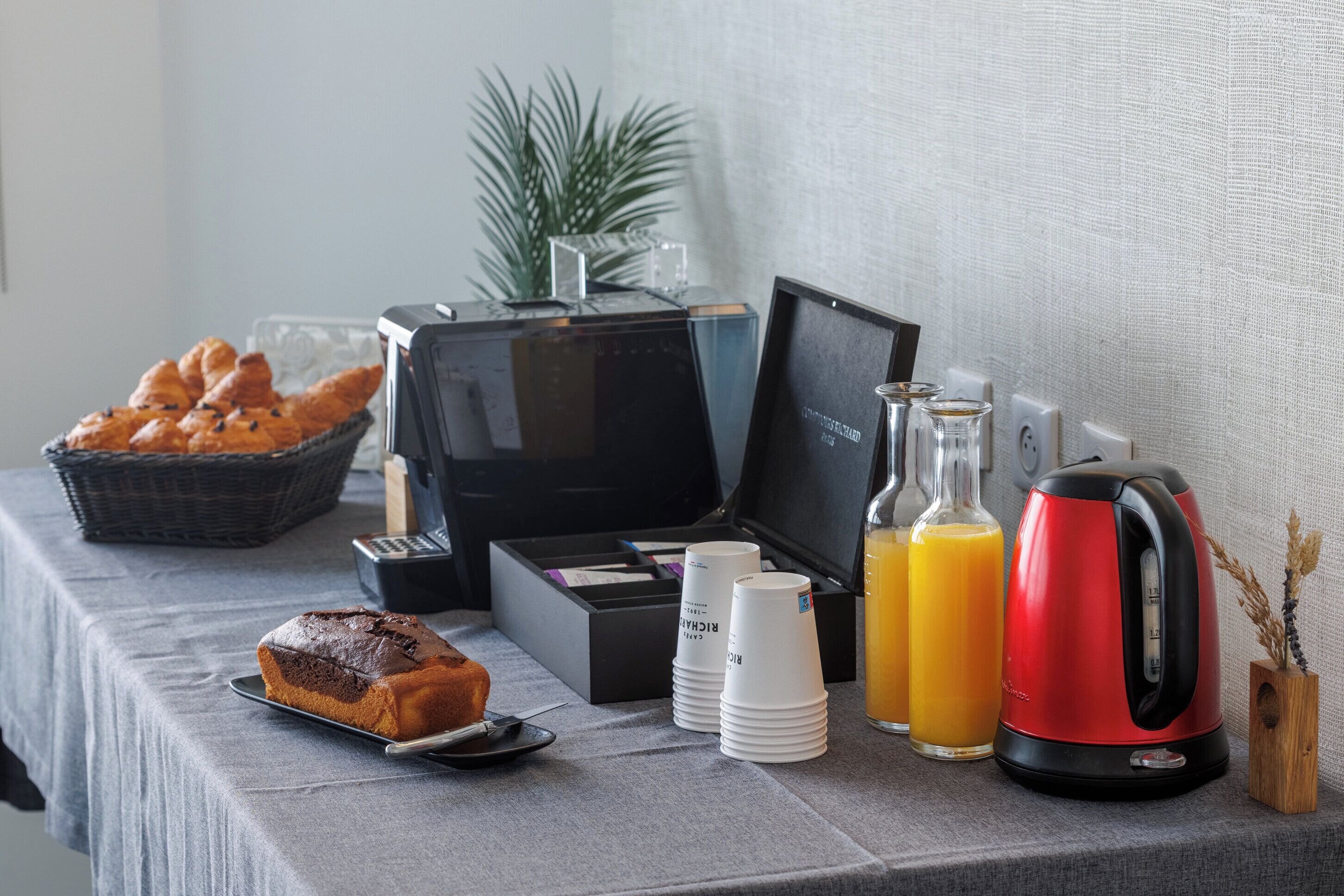 A breakfast setup with croissants, a loaf of bread, coffee cups, a kettle, and orange juice.