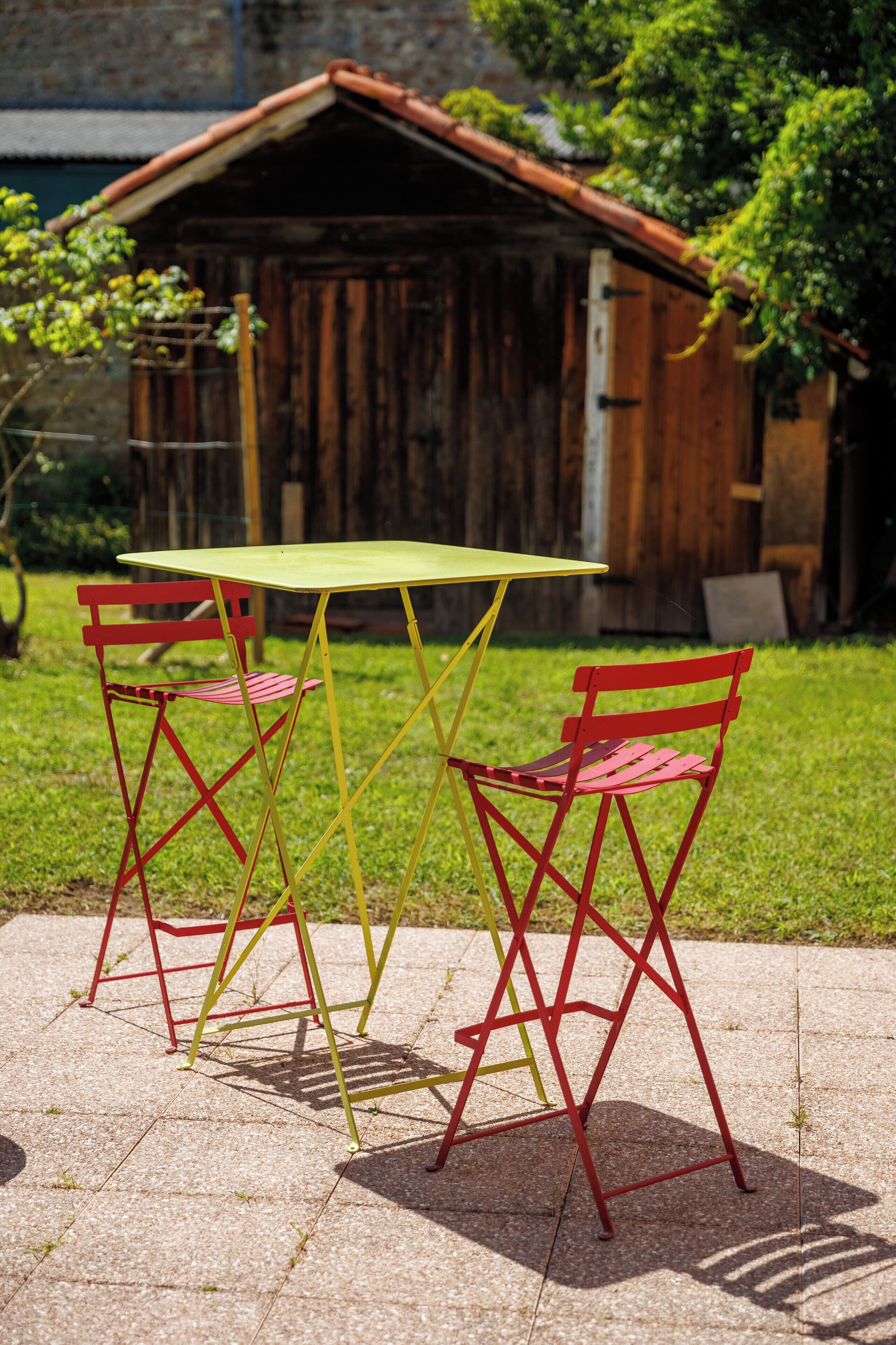 A small outdoor seating area with a table and two chairs, a wooden structure in the background, and a grassy area.