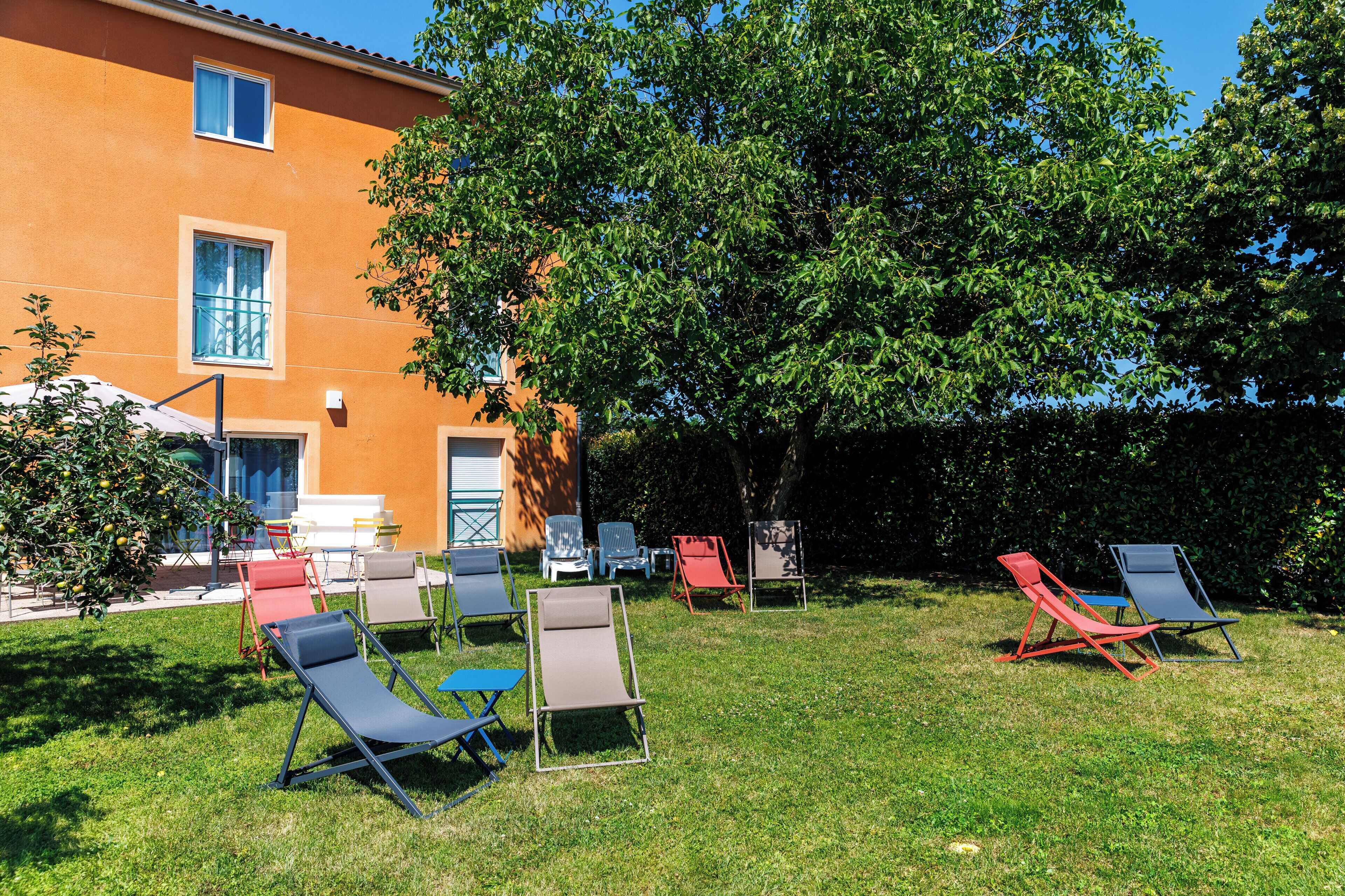 Un espace extérieur aménagé avec plusieurs fauteuils et tables, entouré d’une pelouse bien entretenue et d’un grand arbre.