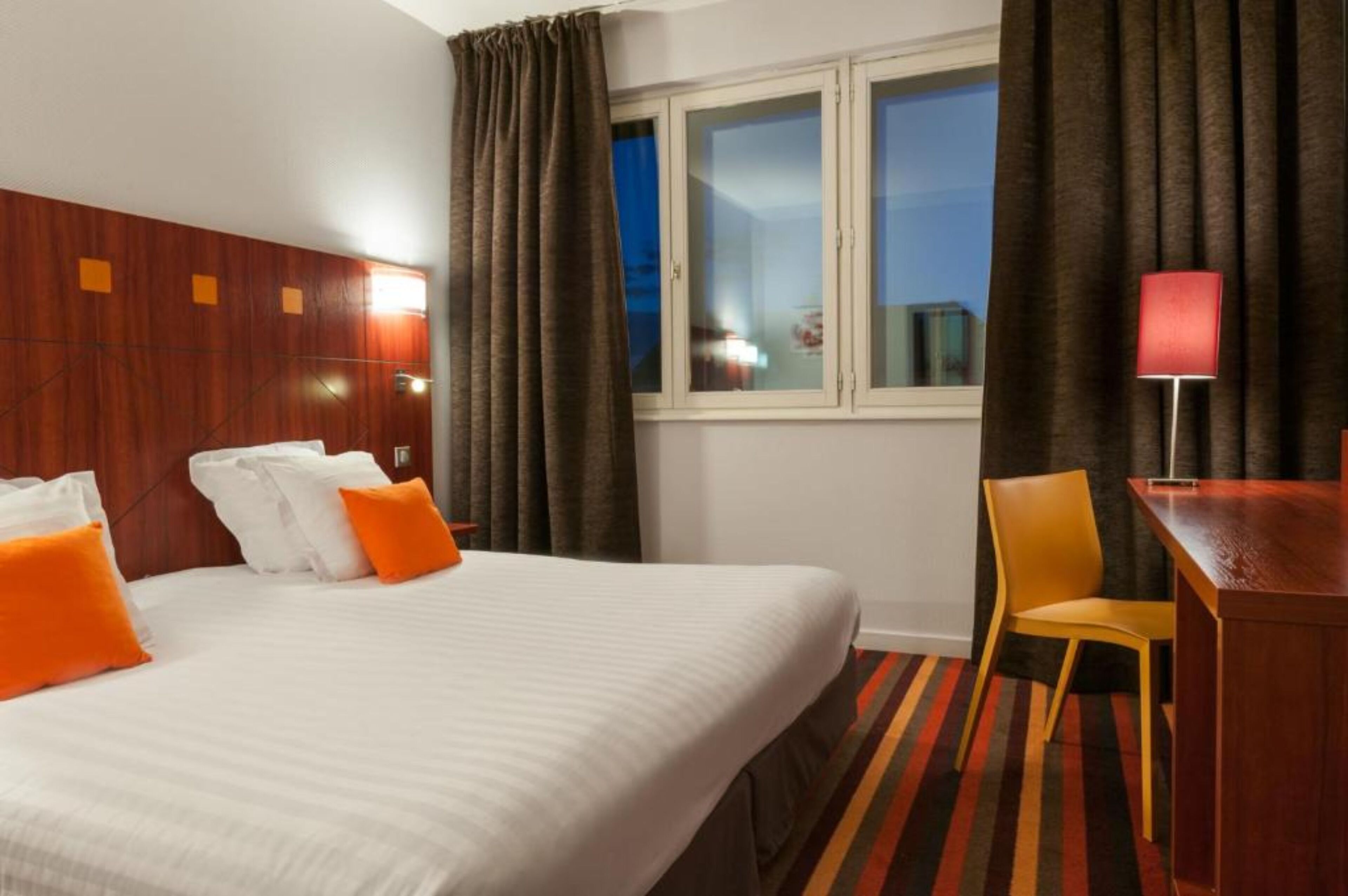 A hotel room with a large bed, a desk, a chair, and a window with curtains.