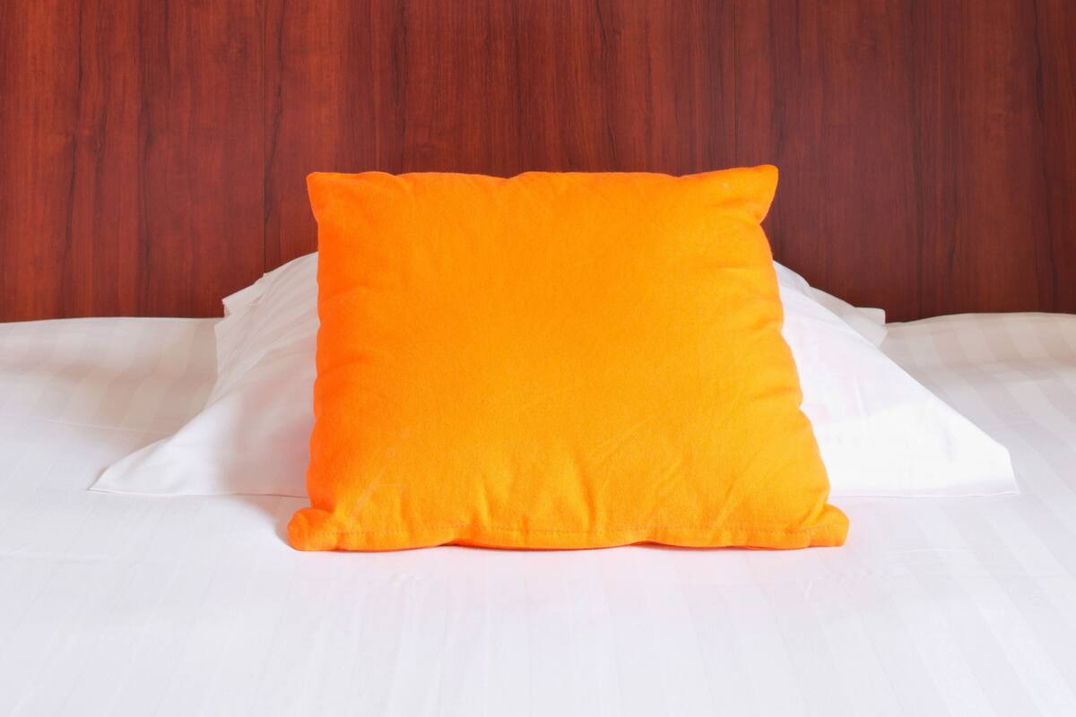 A bed with white sheets and a single bright orange pillow against a wooden headboard with three square cutouts.
