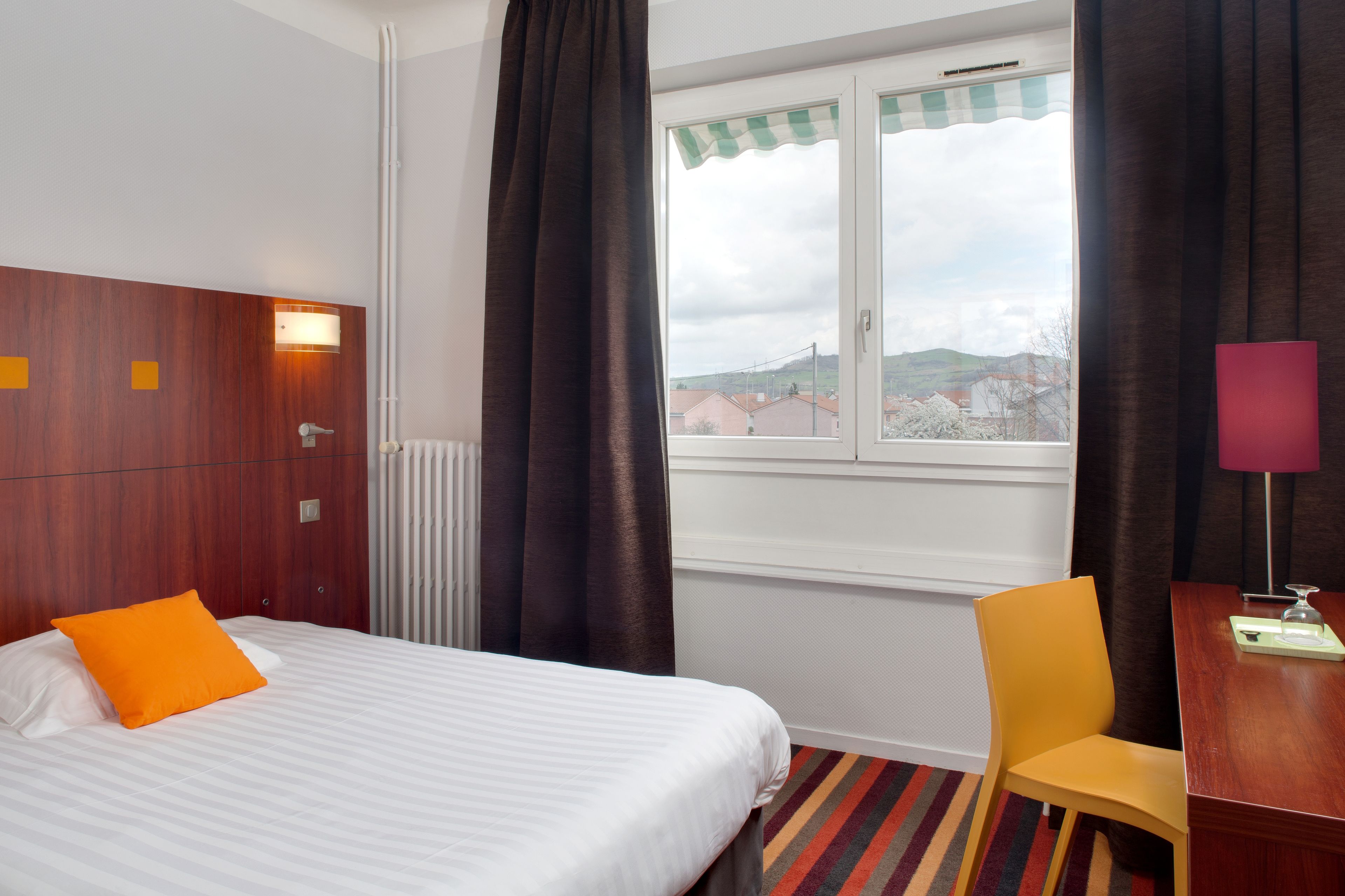 Habitación de hotel con cama, escritorio, silla, ventana con cortinas y vistas a edificios.