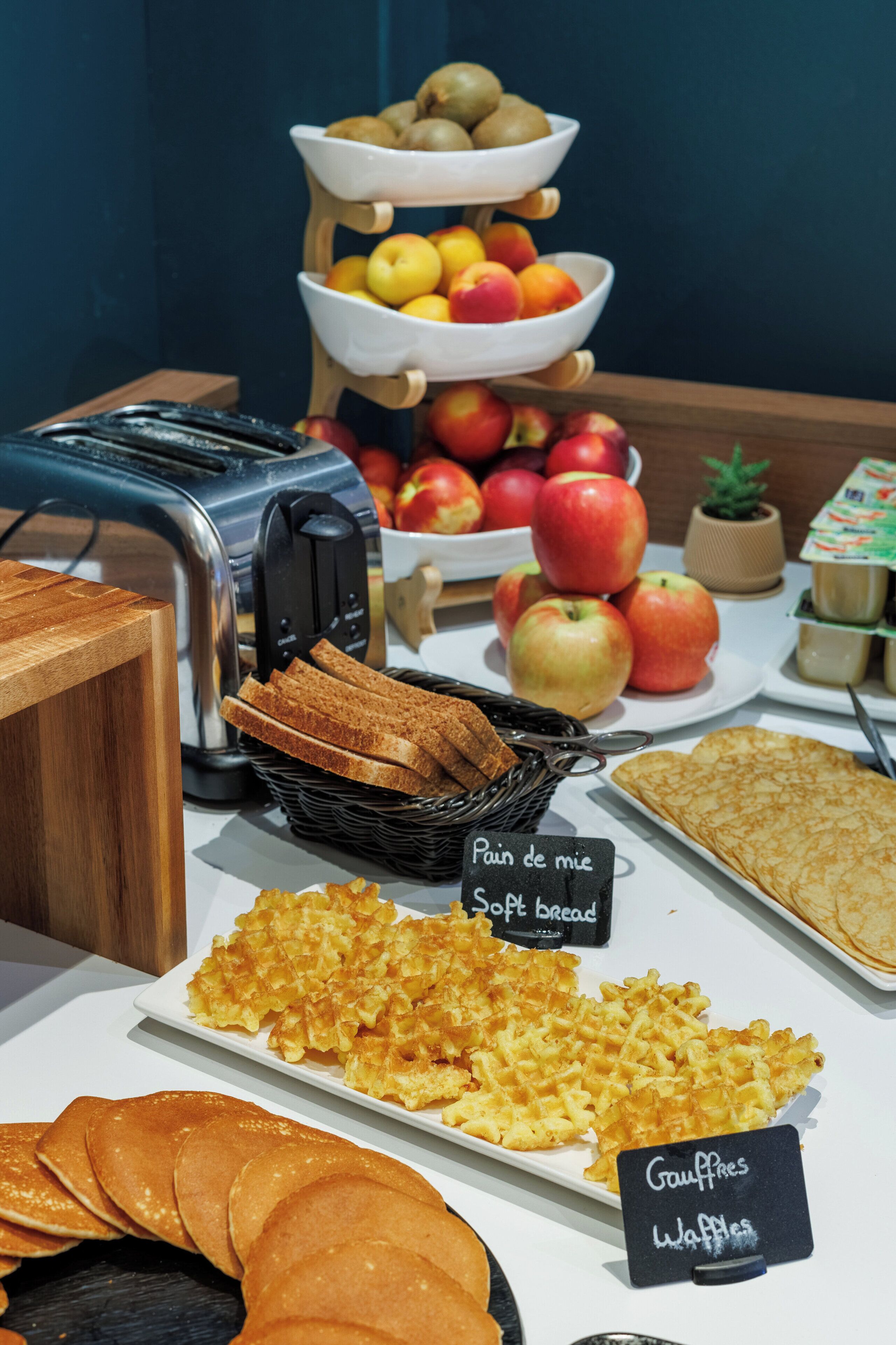 A breakfast buffet with waffles, toast, and a variety of fruits including apples and kiwis.