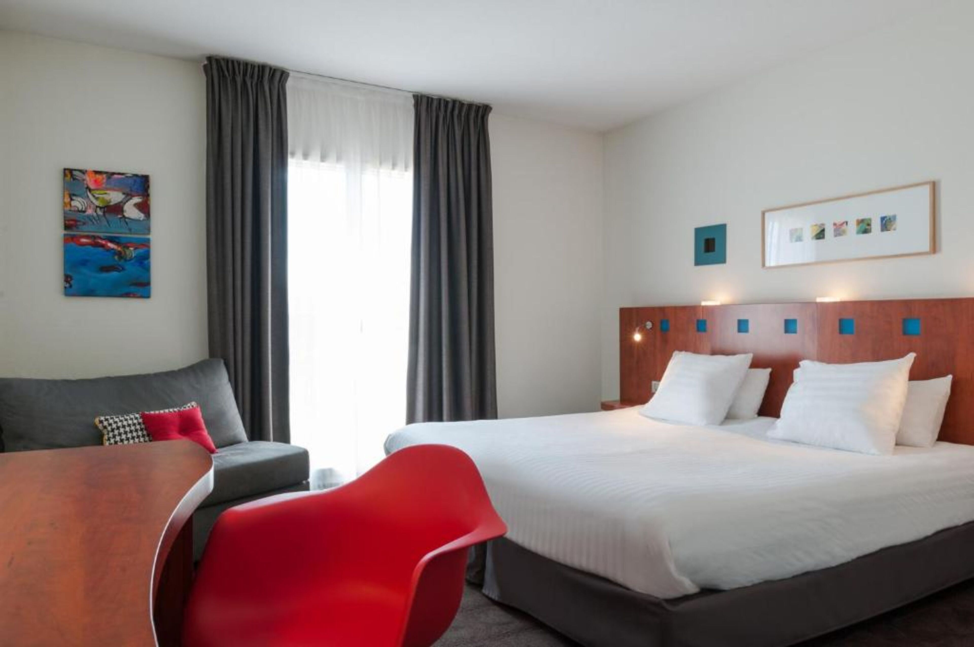 Une chambre d’hôtel avec un lit, une chaise rouge, un canapé gris, une table en bois et un tableau au mur.