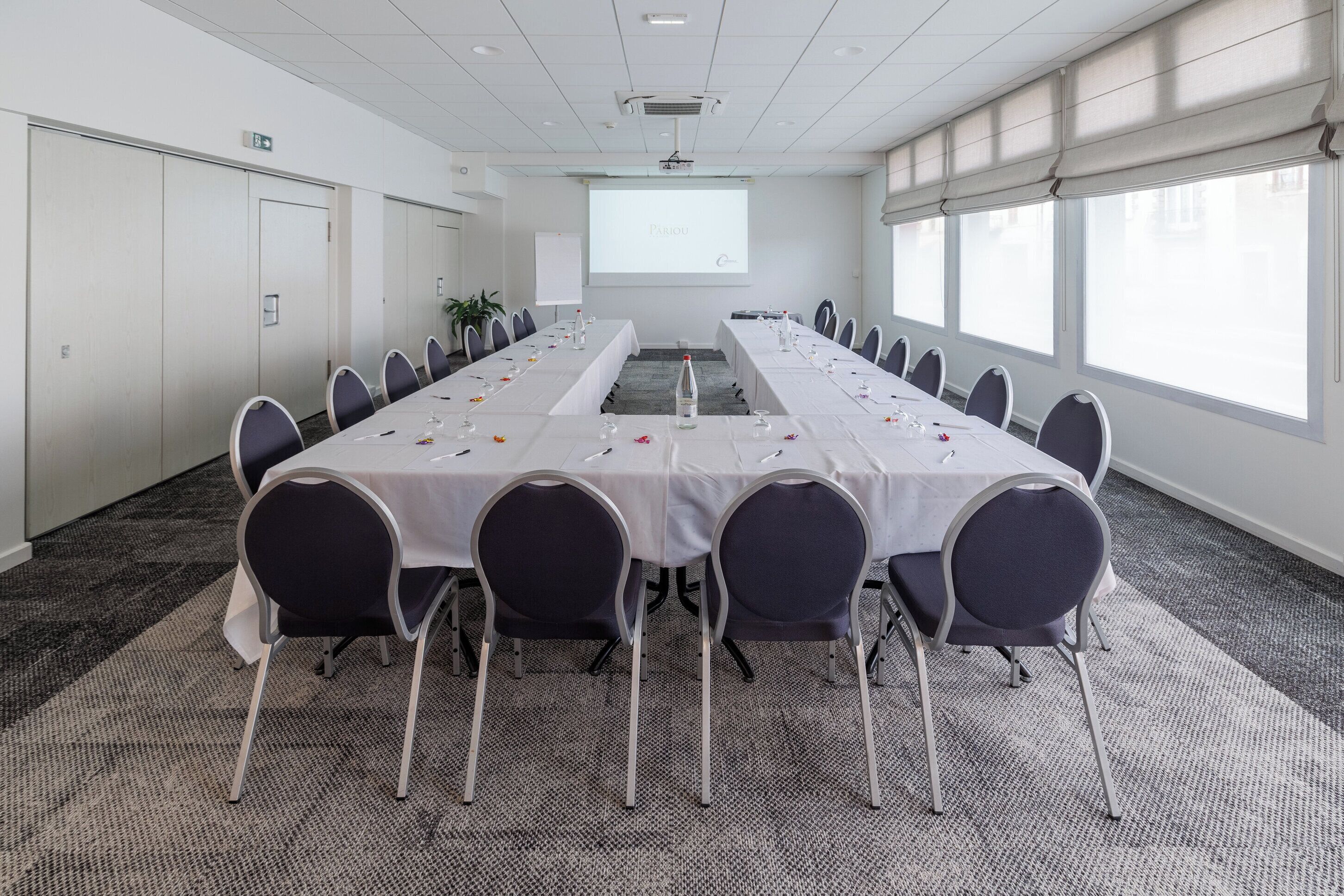 A conference room with a long table, chairs, a projector screen, and windows.