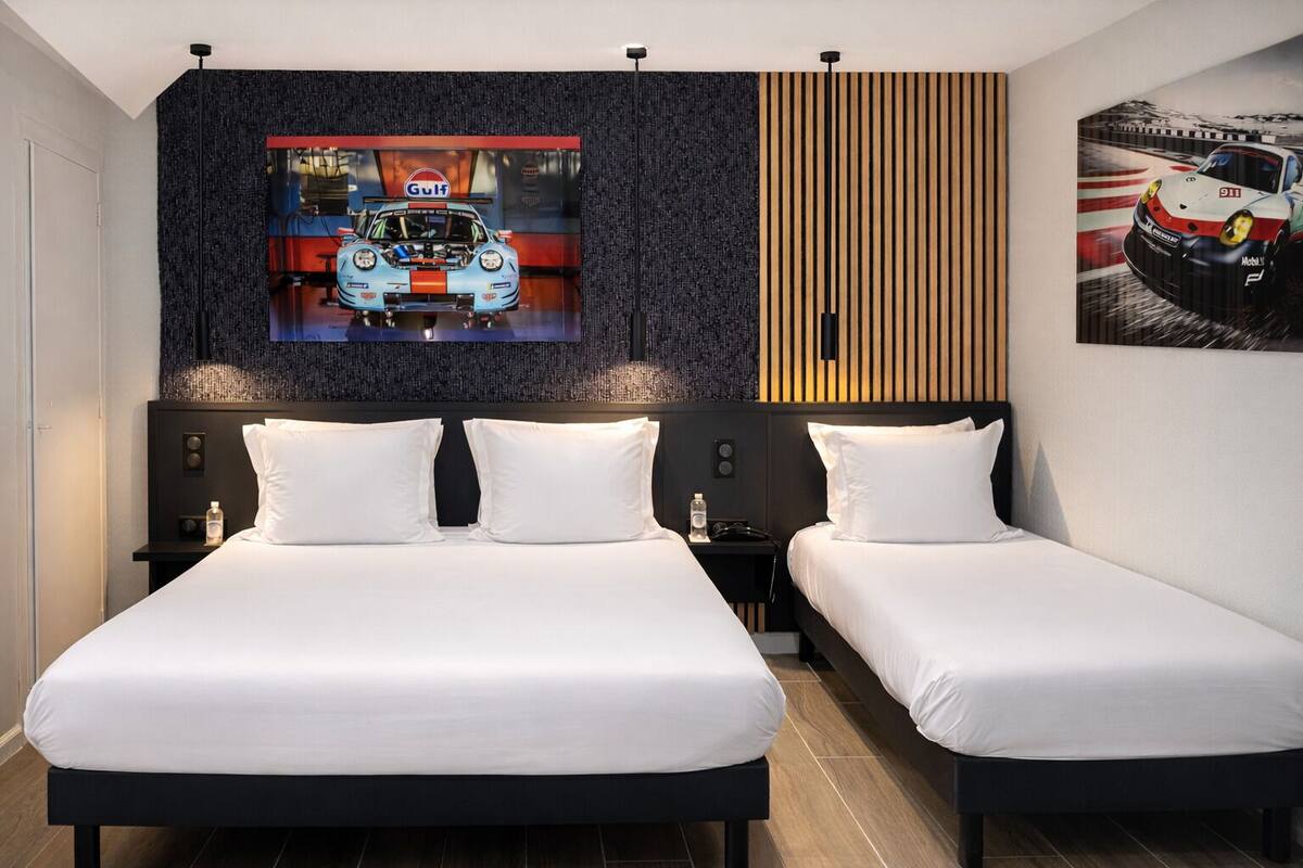 Ein Hotelzimmer mit zwei Betten, einem großen, an der Wand befestigten Fernseher und einem gerahmten Bild von einem Rennwagen.