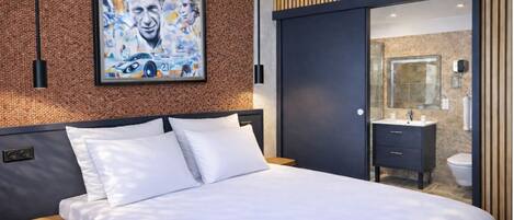 A modern hotel room with a large bed, a wooden headboard, a framed poster on the wall, and a sliding door leading to a bathroom.