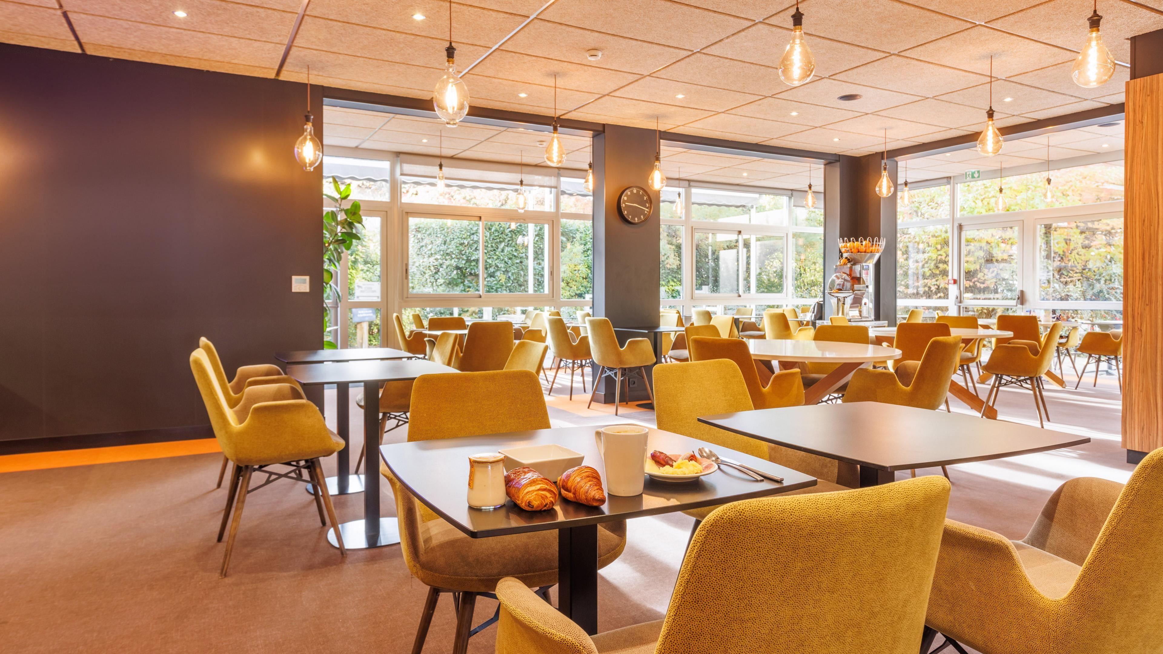 A spacious dining area with yellow chairs and tables, large windows, and hanging light fixtures.