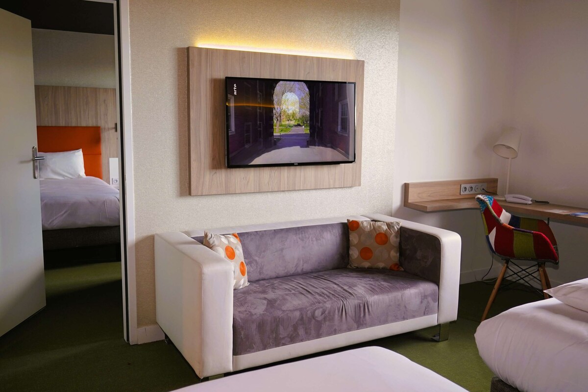 Habitación de hotel moderna con sofá, escritorio, televisor montado en la pared y una cama al fondo.