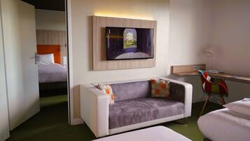A modern hotel room with a sofa, a desk, a TV mounted on the wall, and a bed in the background.