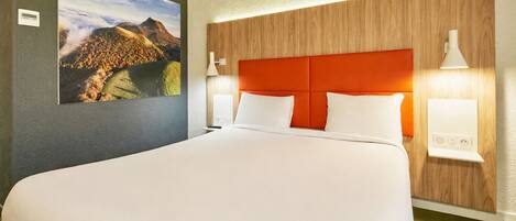 Una habitación de hotel con una cama grande, un cabecero de madera, dos lámparas de noche y un cuadro con un paisaje en la pared.