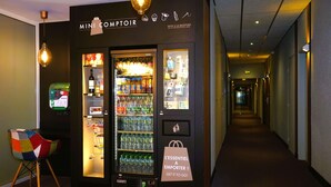 A Mini Comptoir vending machine with a variety of beverages and snacks, located in a hallway with a carpeted floor and a colorful chair. - Kyriad Clermont Ferrand Nord - Riom (Riom)