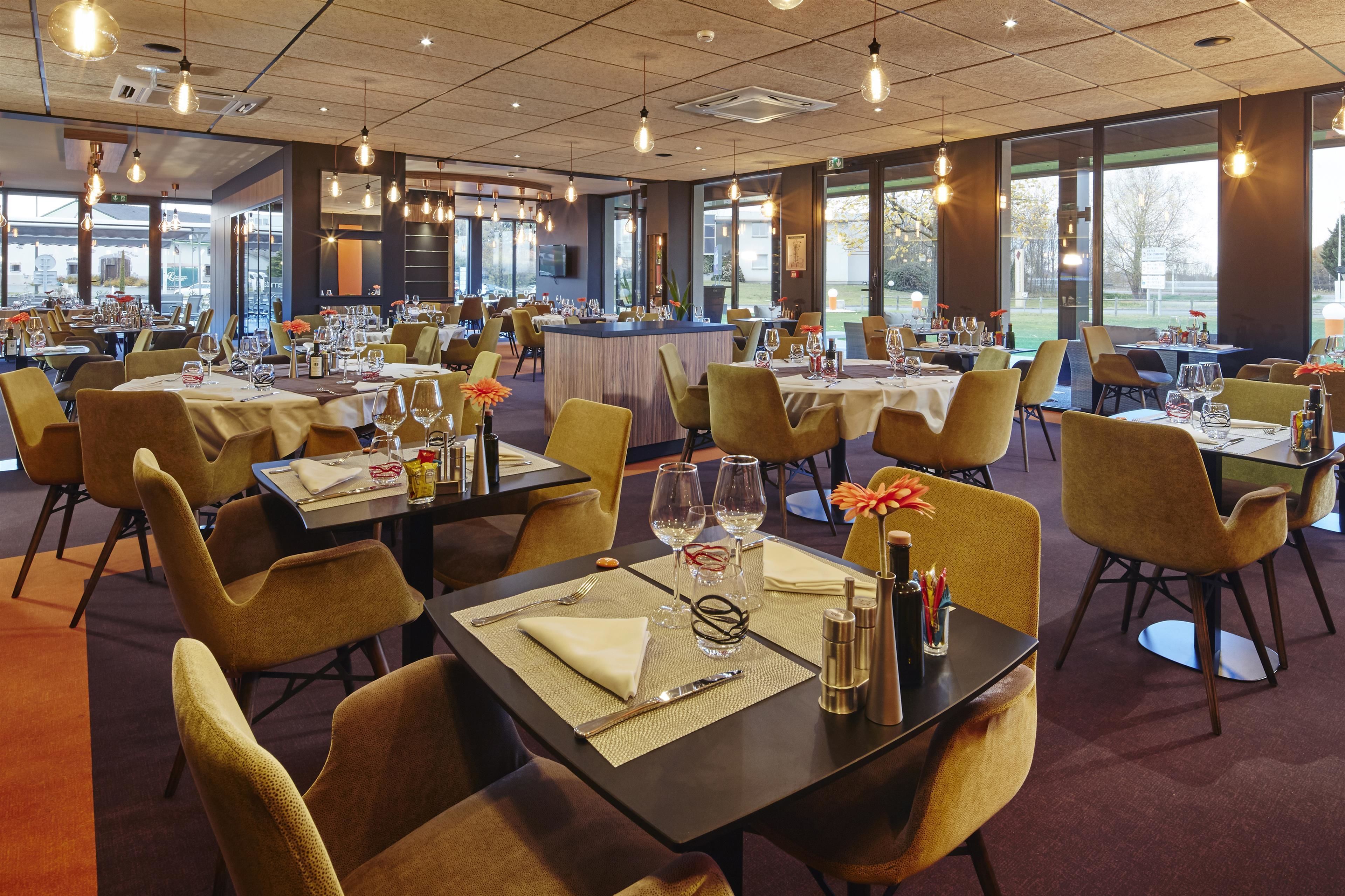 A spacious dining area with multiple tables set for dining, a central bar area, and large windows providing natural light and views of the outdoors.