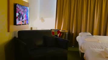 A hotel room with a sofa, a bed with white linens, a television mounted on the wall, a lamp, and a curtain.
