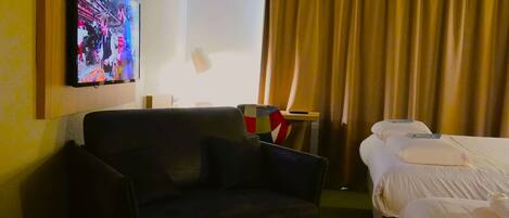 A hotel room with a sofa, a bed with white linens, a television mounted on the wall, a lamp, and a curtain.