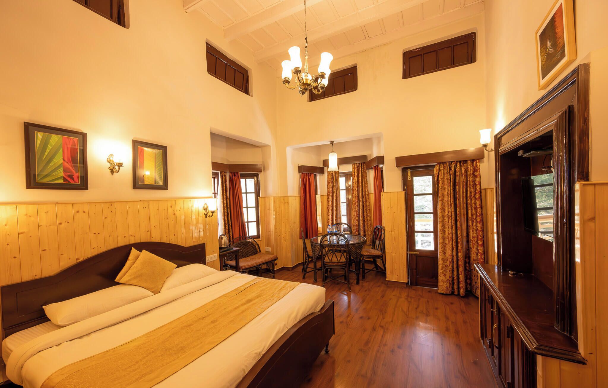 A hotel room with a large bed, a dining table with chairs, a TV, and a chandelier.