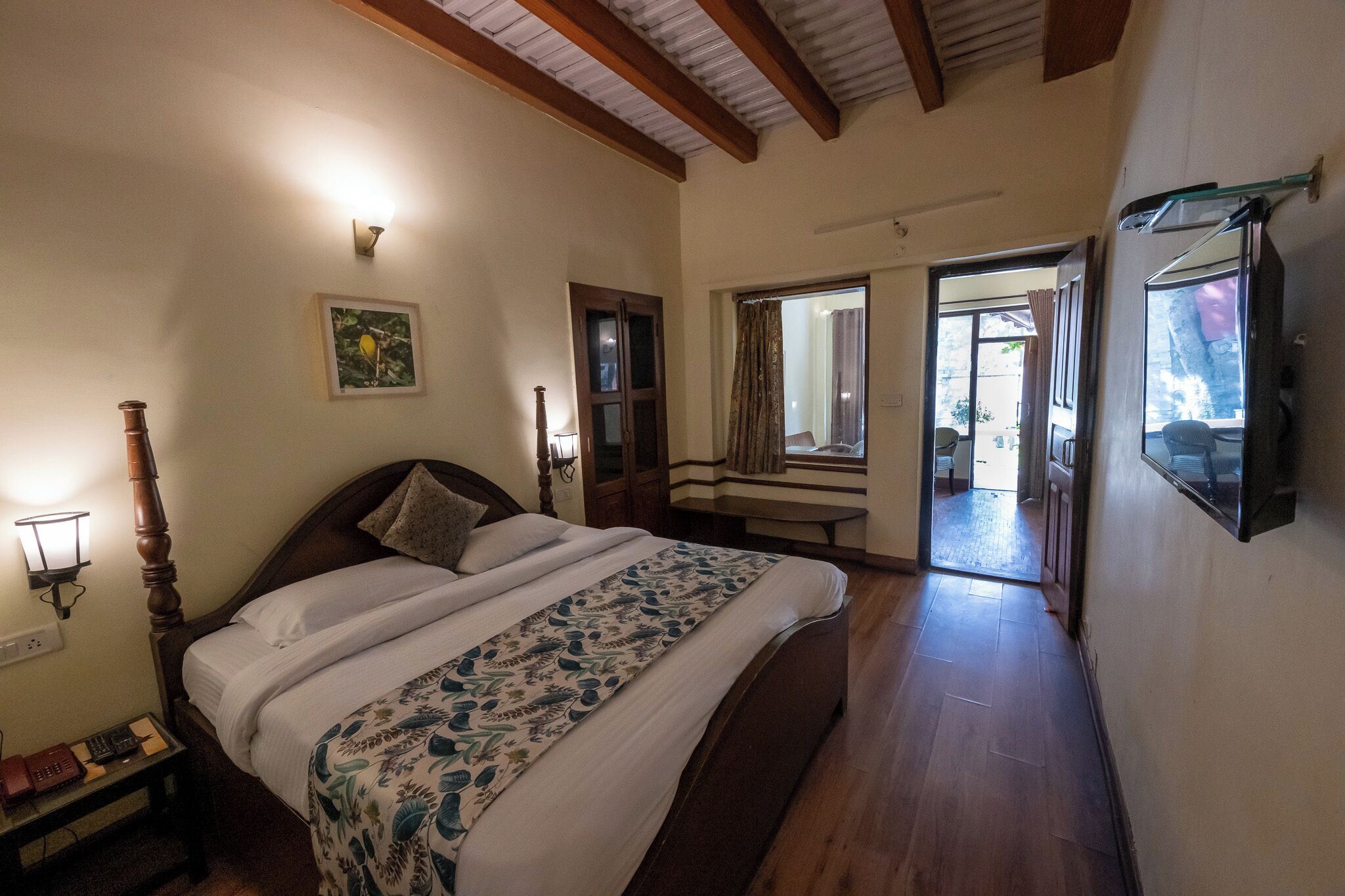 A bedroom with a wooden bed, a bedside table with a lamp, a framed picture on the wall, and a window with curtains.