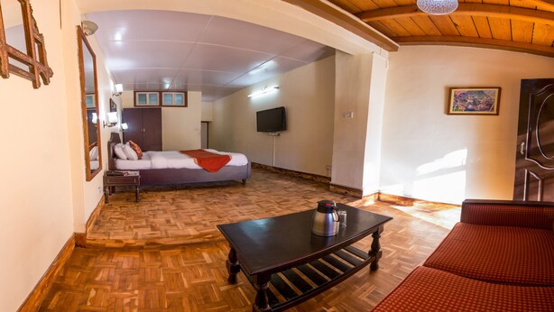A room with a wooden herringbone floor, a bed with a red blanket, a black table with a kettle, a red sofa, and a television on the wall.