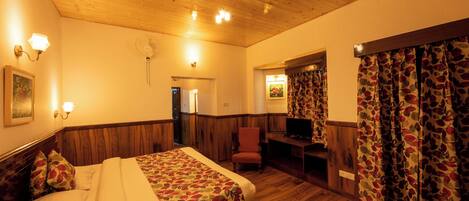 A hotel room with a wooden ceiling, a bed with floral bedding, a chair, a desk with a television, and a window with floral curtains.