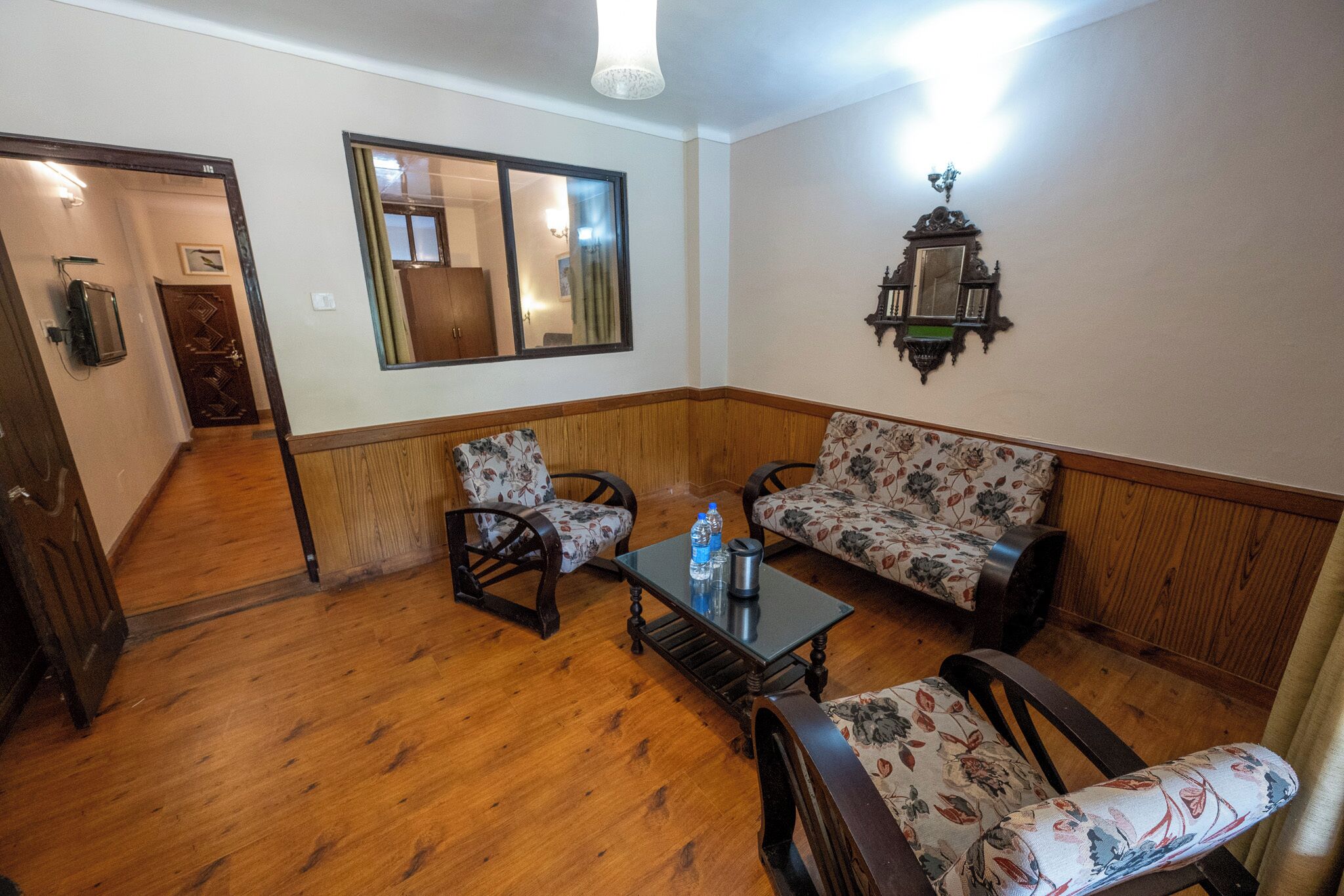 Ein Zimmer mit Holzboden, einer Couch, zwei Sesseln, einem kleinen Tisch, einem Spiegel und einer Wandlampe.