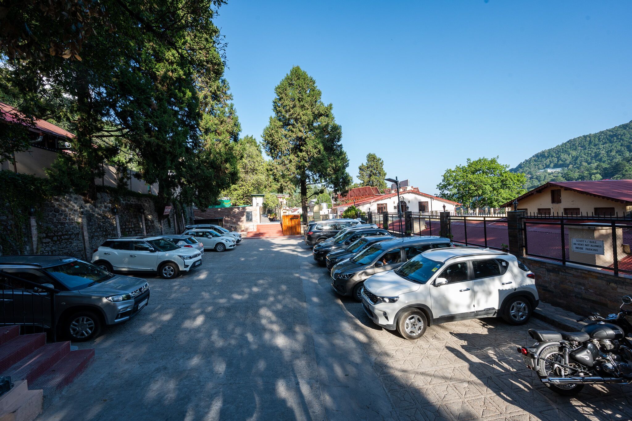 A parking lot with several cars and a motorcycle, surrounded by trees and buildings with red roofs.