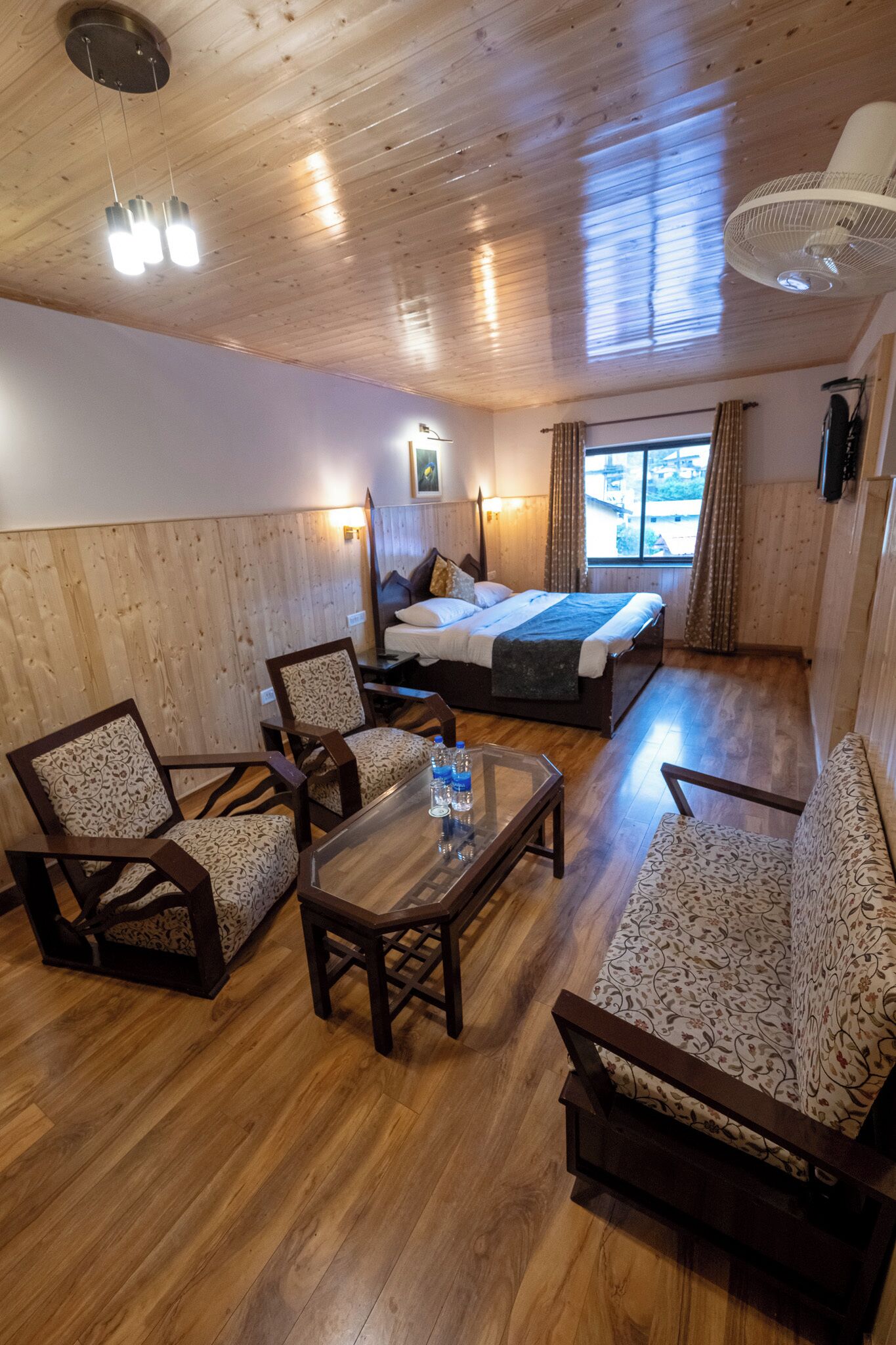 Una habitación acogedora con una cama, dos sillones estampados, una mesita con una botella, suelos de madera, un ventilador de techo y una ventana con cortinas.