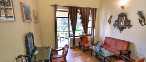 A cozy room with a wooden floor, a sofa, a table, and chairs. There is a window with curtains, a wall-mounted light, and framed pictures.