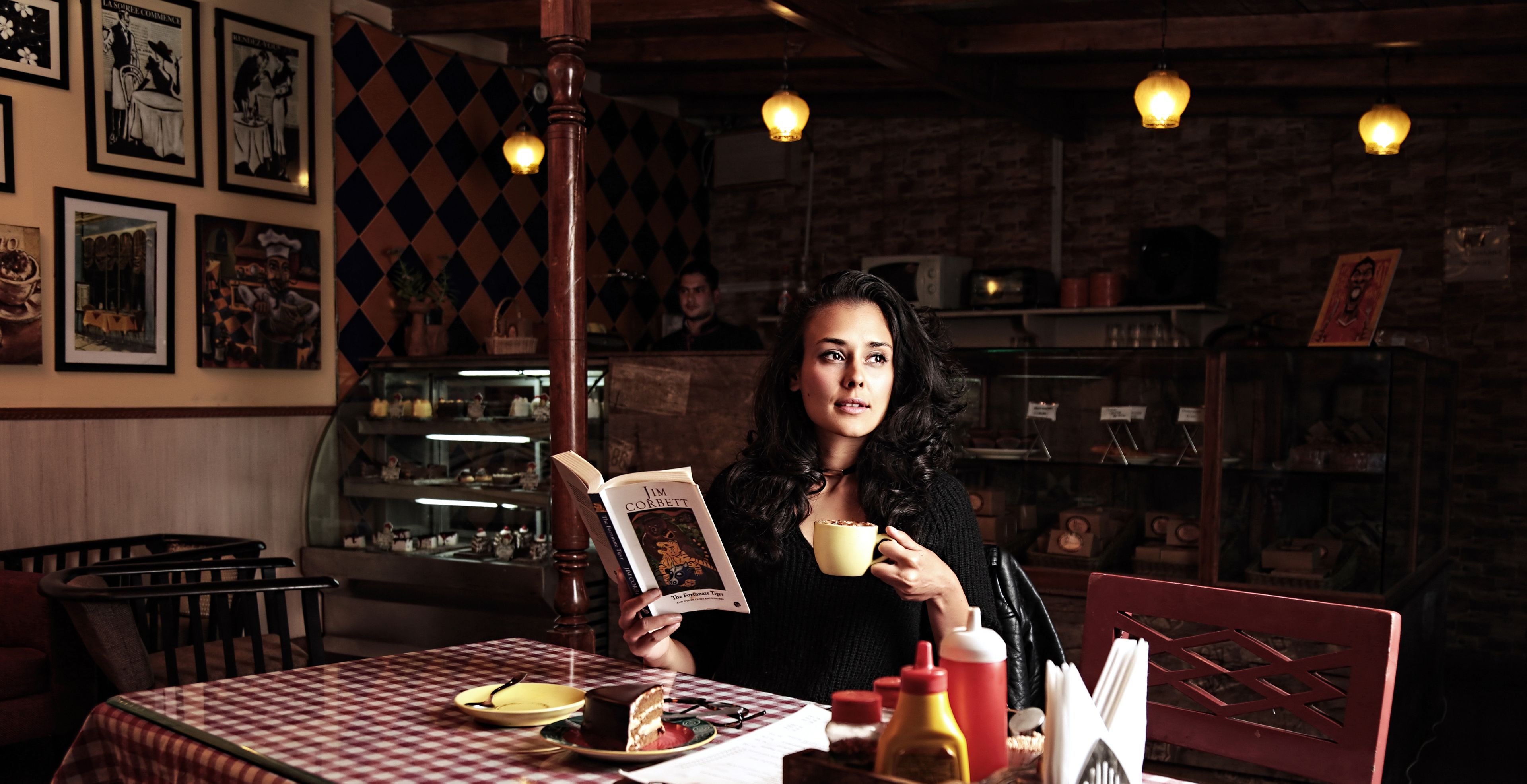 A person reading a book at a table with a checkered tablecloth, a cake, and a cup.