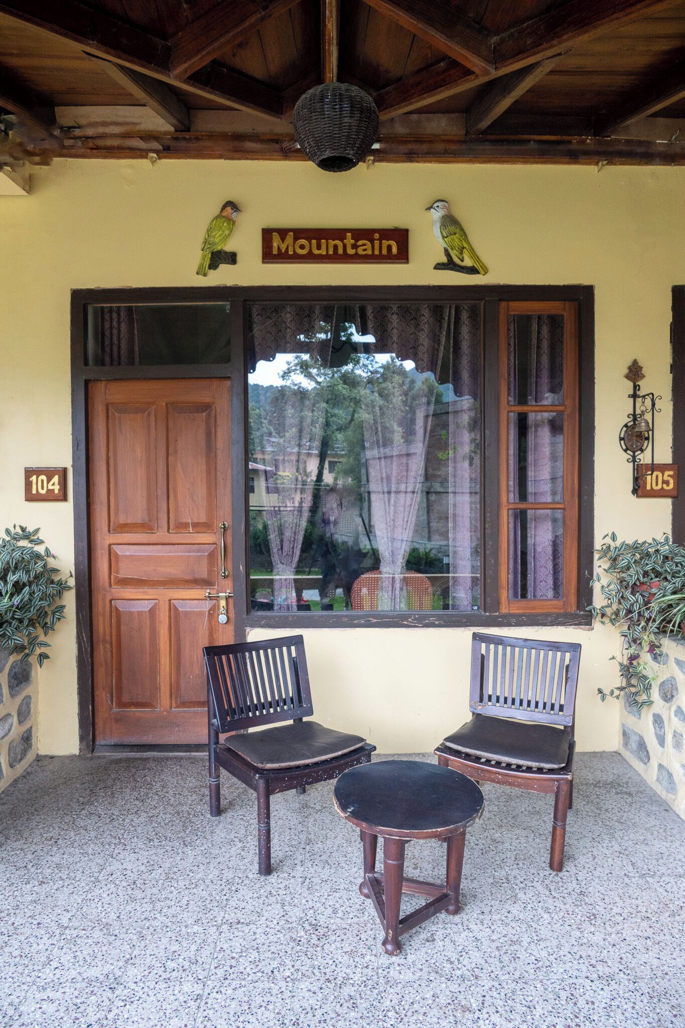 Una puerta de madera con el número 104, una ventana con cortinas, dos sillas de madera y un cartel sobre la ventana que dice "Mountain".