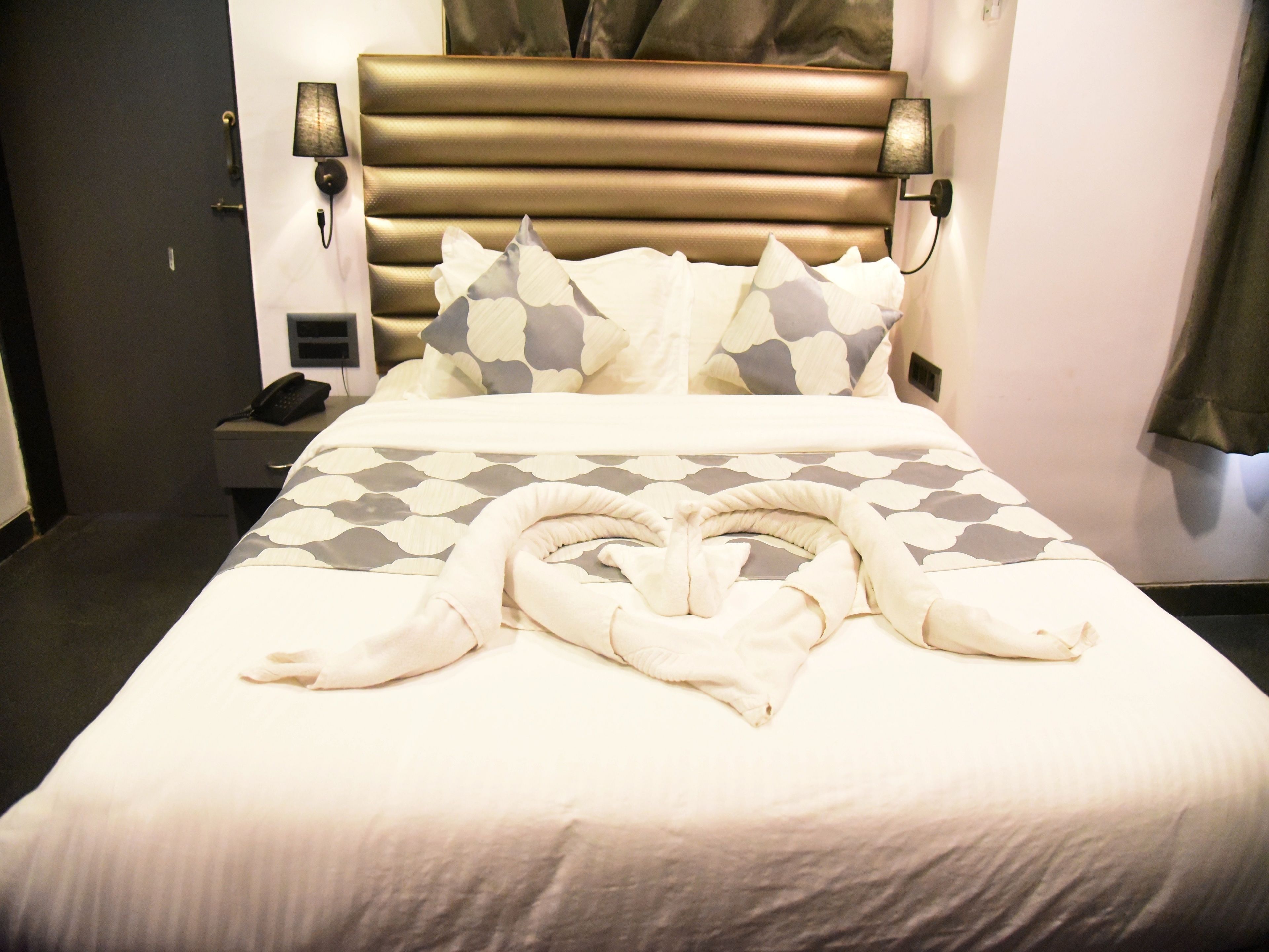 A hotel room with a bed, a bedside table with a phone, and a wall-mounted lamp.