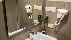 A hotel bathroom with a large mirror, a white sink, and hair dryers mounted on the wall.
