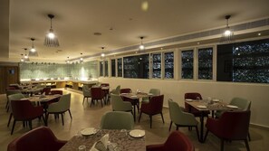 A restaurant with multiple dining tables and chairs, a bar area, and large windows with a night view.