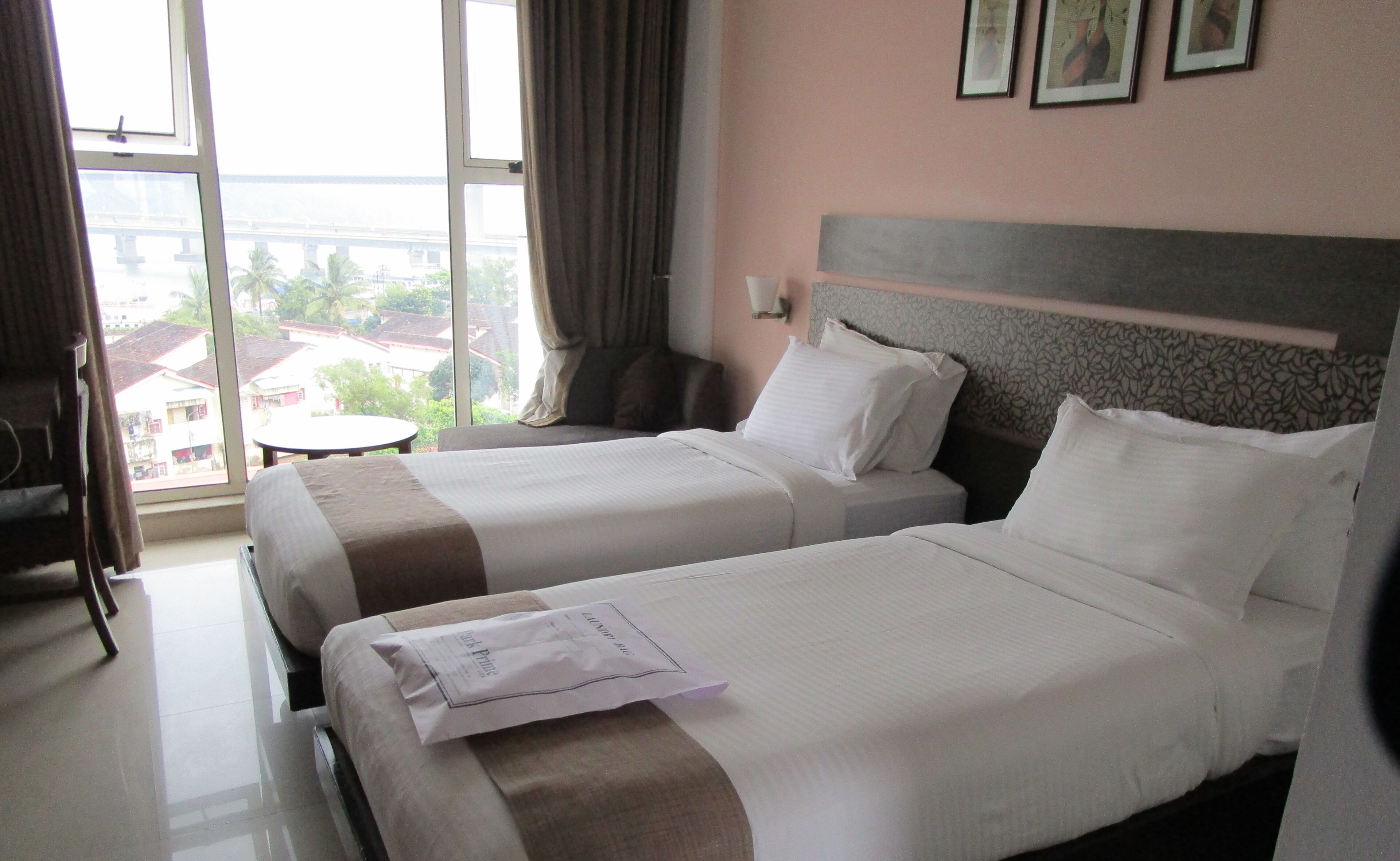 A hotel room with two beds, a small table, and a view of buildings through large windows.