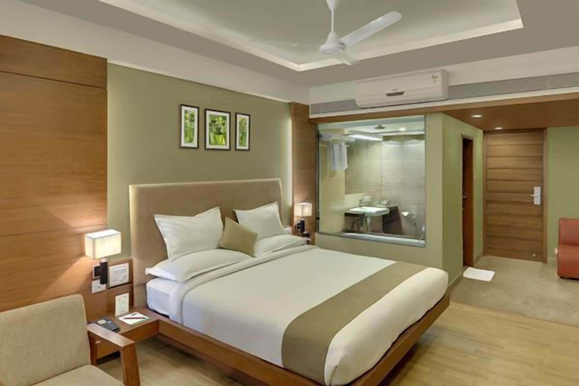 A hotel room with a large bed, a bedside table, a chair, and a bathroom visible through a glass partition.