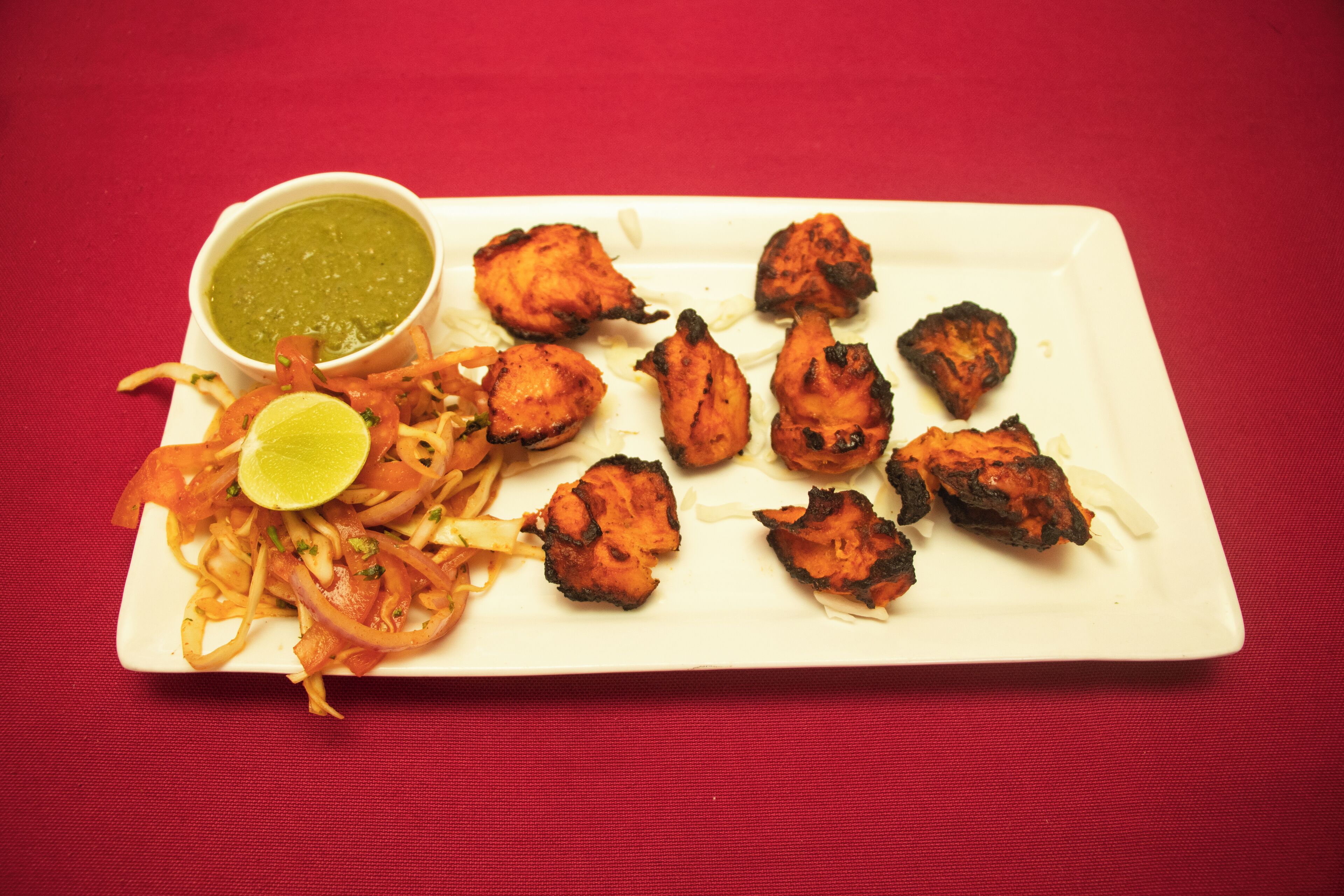 A plate of tandoori chicken with a side of sliced onions and a lime wedge, accompanied by a bowl of green chutney.