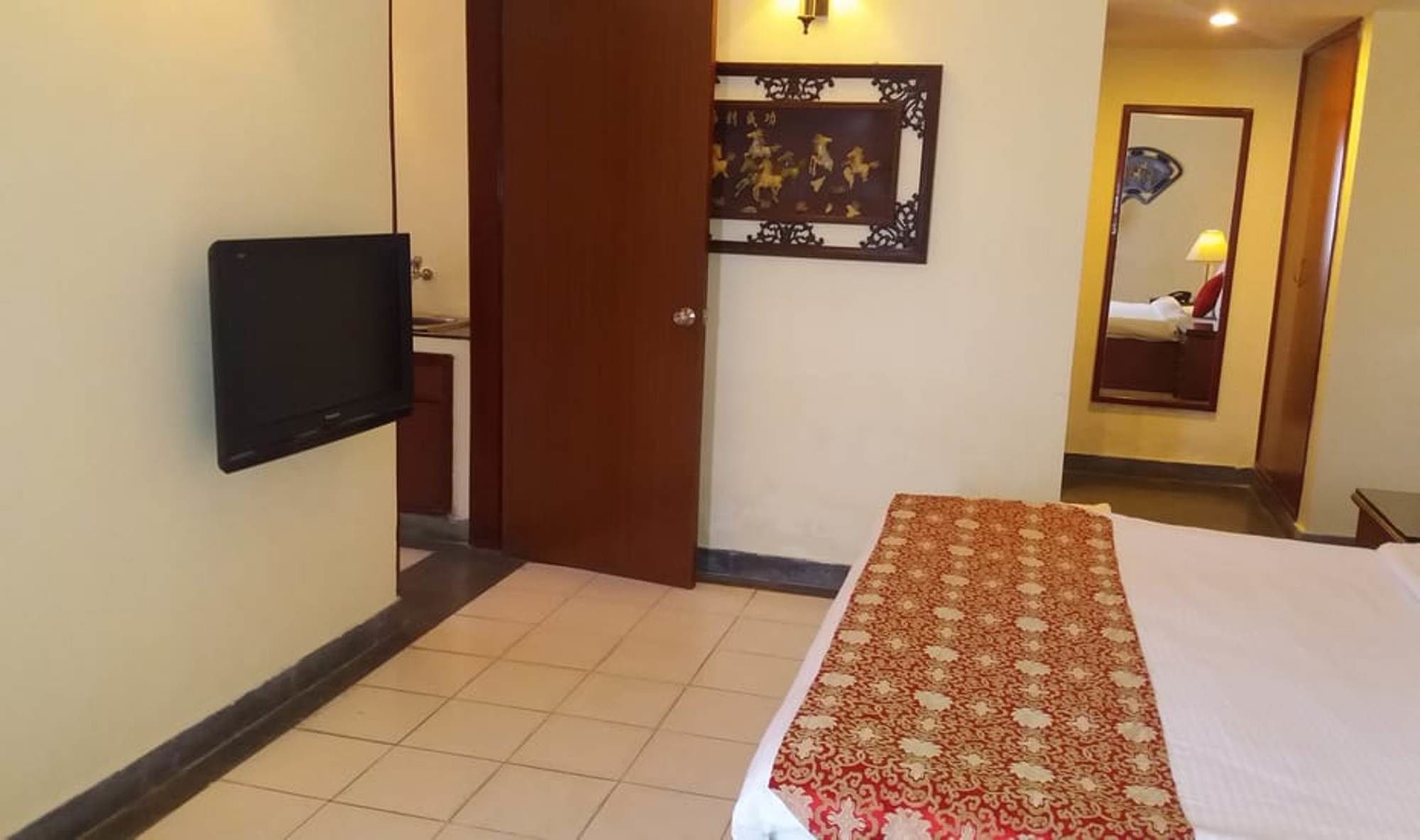 A hotel room with a bed, a flat-screen TV mounted on the wall, a decorative mirror, and a patterned rug.