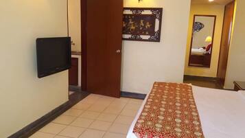 A hotel room with a bed, a flat-screen TV mounted on the wall, a decorative mirror, and a patterned rug.