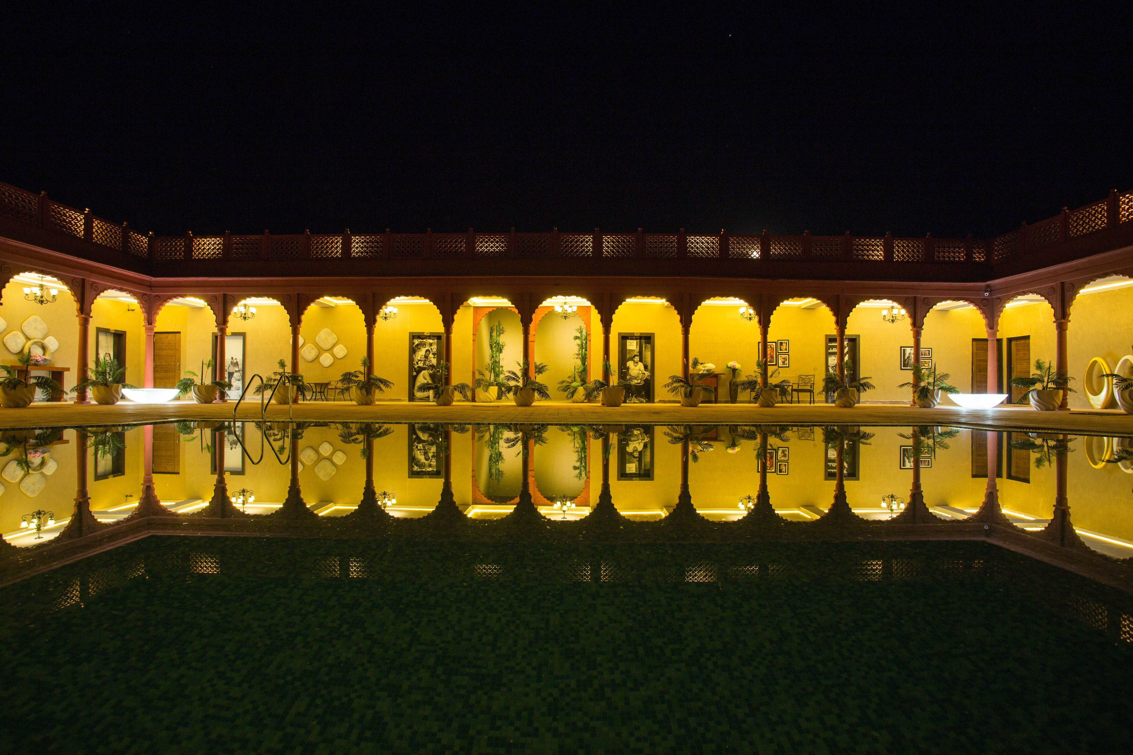 Ein Hotel bei Nacht mit einem spiegelnden Pool, bogenförmigen Balkonen und Kübelpflanzen.