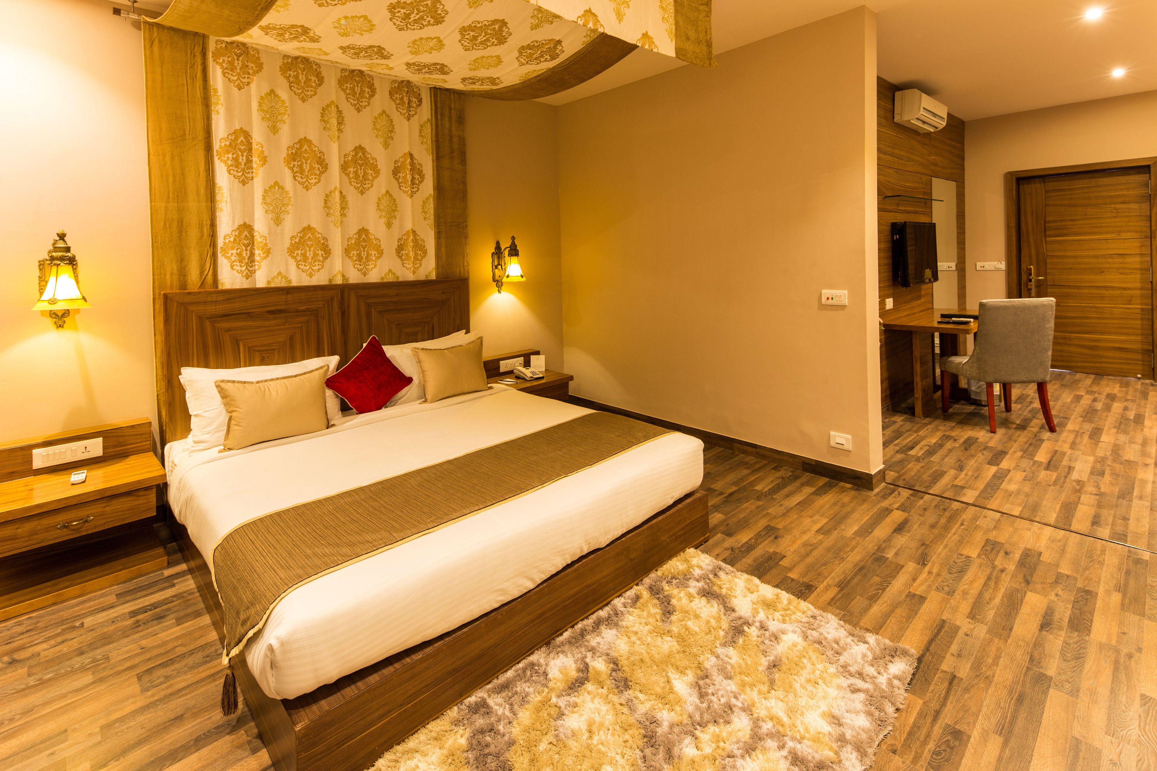 A hotel room with a large bed, two bedside lamps, a desk with a chair, and a wooden floor.