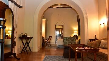 A cozy living room with a wooden floor, a patterned sofa, a coffee table, and a dining table with chairs. An arched doorway leads to another room.