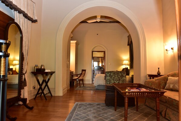 A cozy living room with a wooden floor, a patterned sofa, a coffee table, and a dining table with chairs. An arched doorway leads to another room.