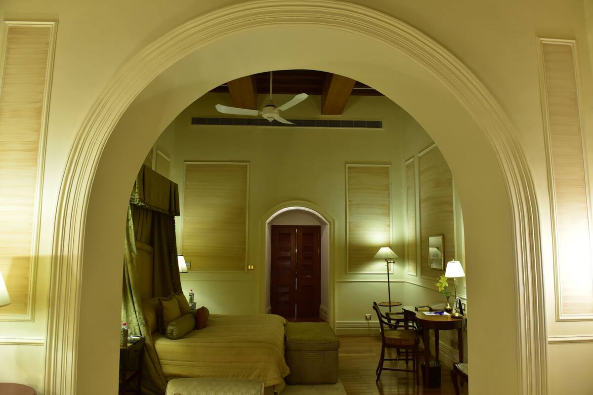Ein Hotelzimmer mit einem großen Bett, einem Schreibtisch, einem Stuhl, einem Deckenventilator und einem Rundbogenportal.