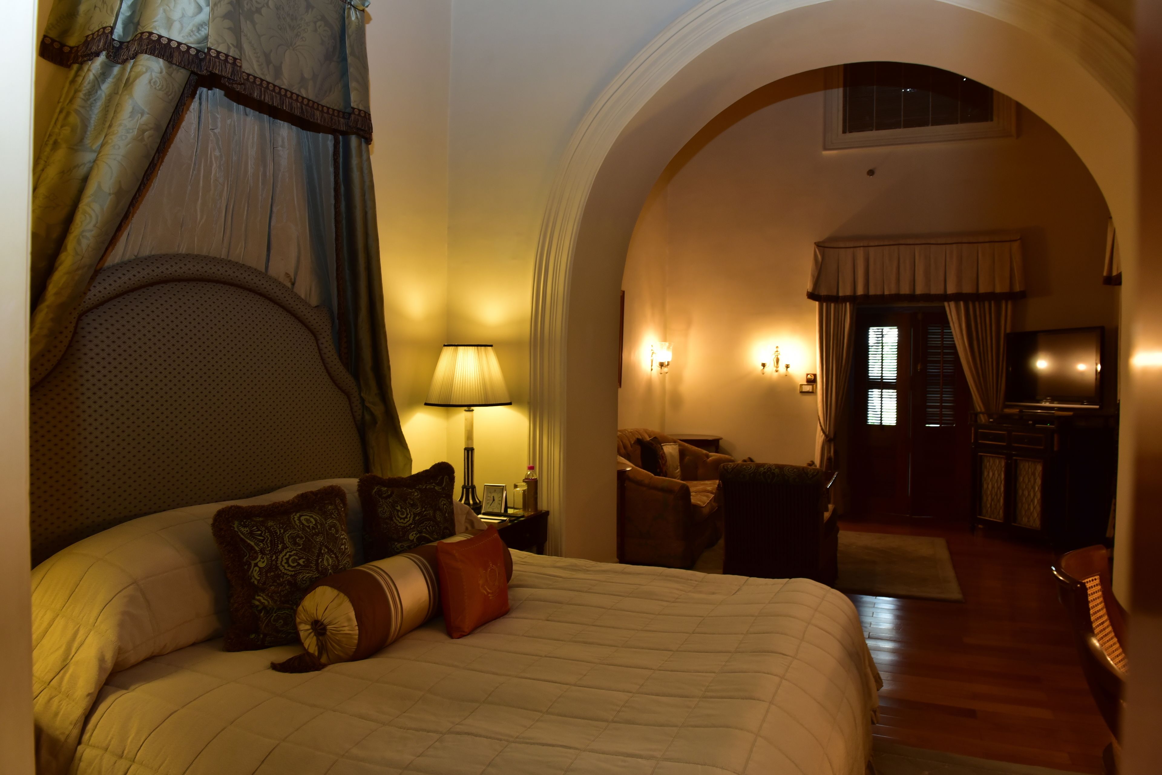 A hotel room with a large bed, a nightstand, a lamp, and a view through an arched doorway to another room.