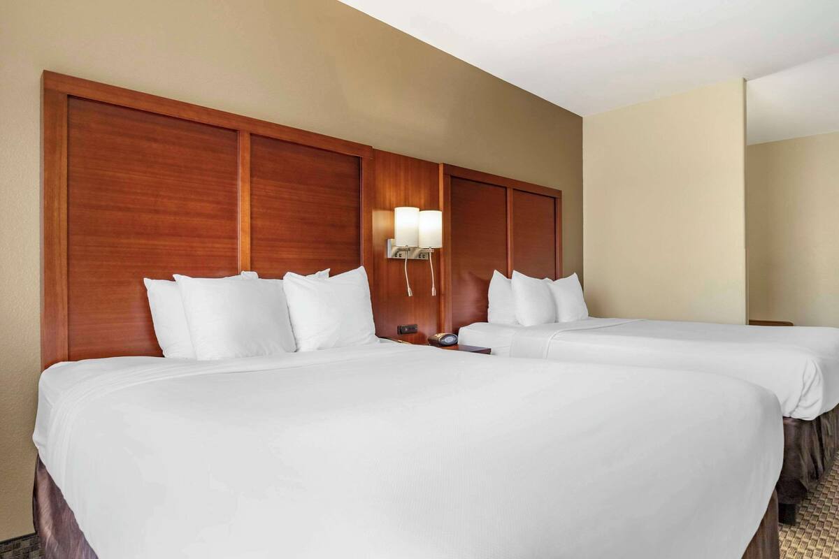 Une chambre d’hôtel avec deux lits, une tête de lit en bois, du linge de lit blanc et un luminaire fixé au mur.