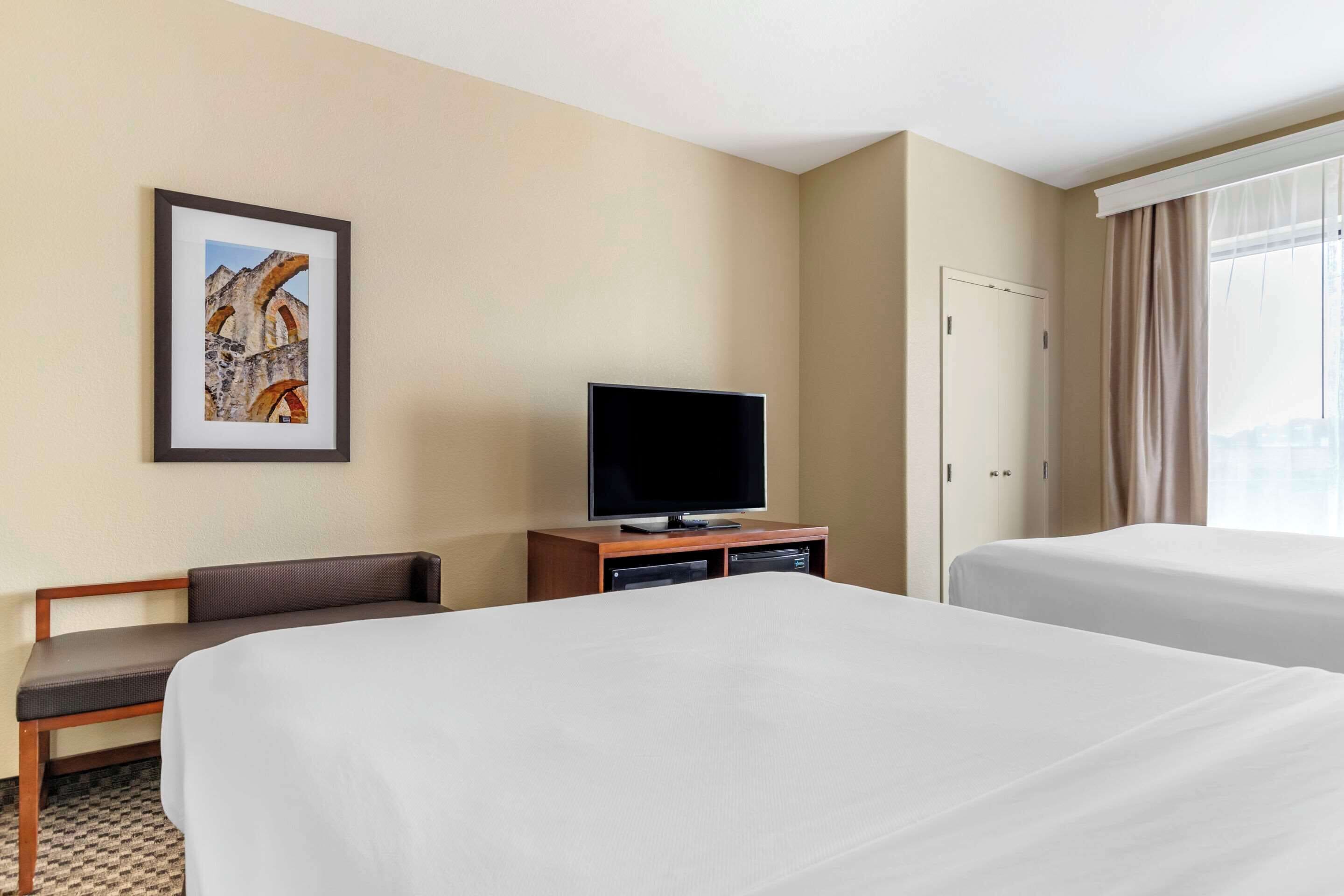 A hotel room with two beds, a television on a wooden stand, a framed picture on the wall, and a window with curtains.